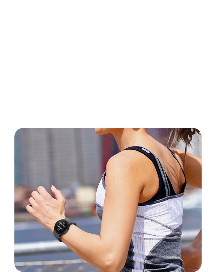 A woman's wrist is shown in the middle of running in the city. She is wearing a black Galaxy Watch FE, which displays the time, the duration of exercise, the calories burned, and her heart rate.