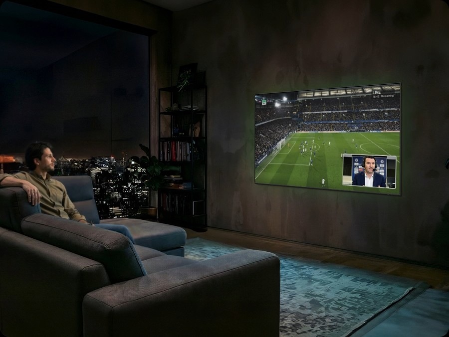 Man watches football on Samsung TV at night. Soundwaves emanate from TV's side and front. He speaks into remote control ‘Bixby, AI Football Mode ON’. AI Football Mode turns on, soundwaves show balanced crowd and commentator sound, and the field looks more crisp and vibrant than when it's off.