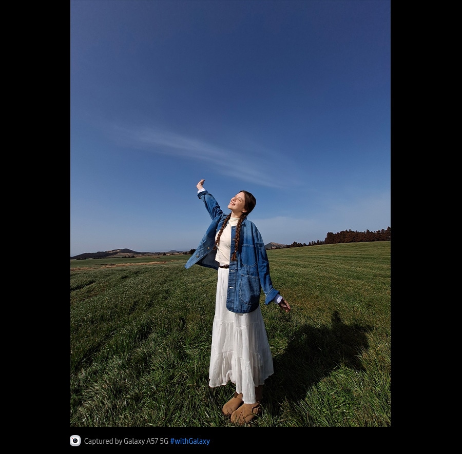 Photo of a young woman standing in the middle of an open grass field against the blue sky, taken with Galaxy A57 5G's Ultra Wide Camera. Photo details are bright and vivid with improved quality.
