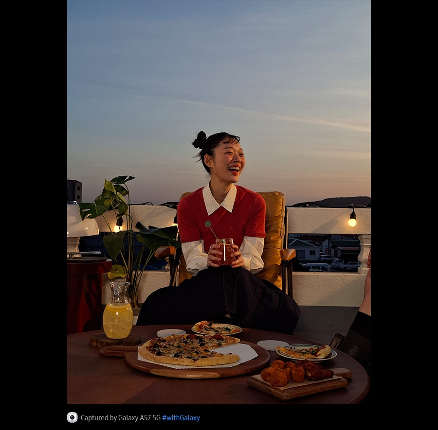 Photo of a young woman smiling on an outdoor terrace taken in a low-lit environment with Galaxy A57 5G. Photo details are bright and crisp.