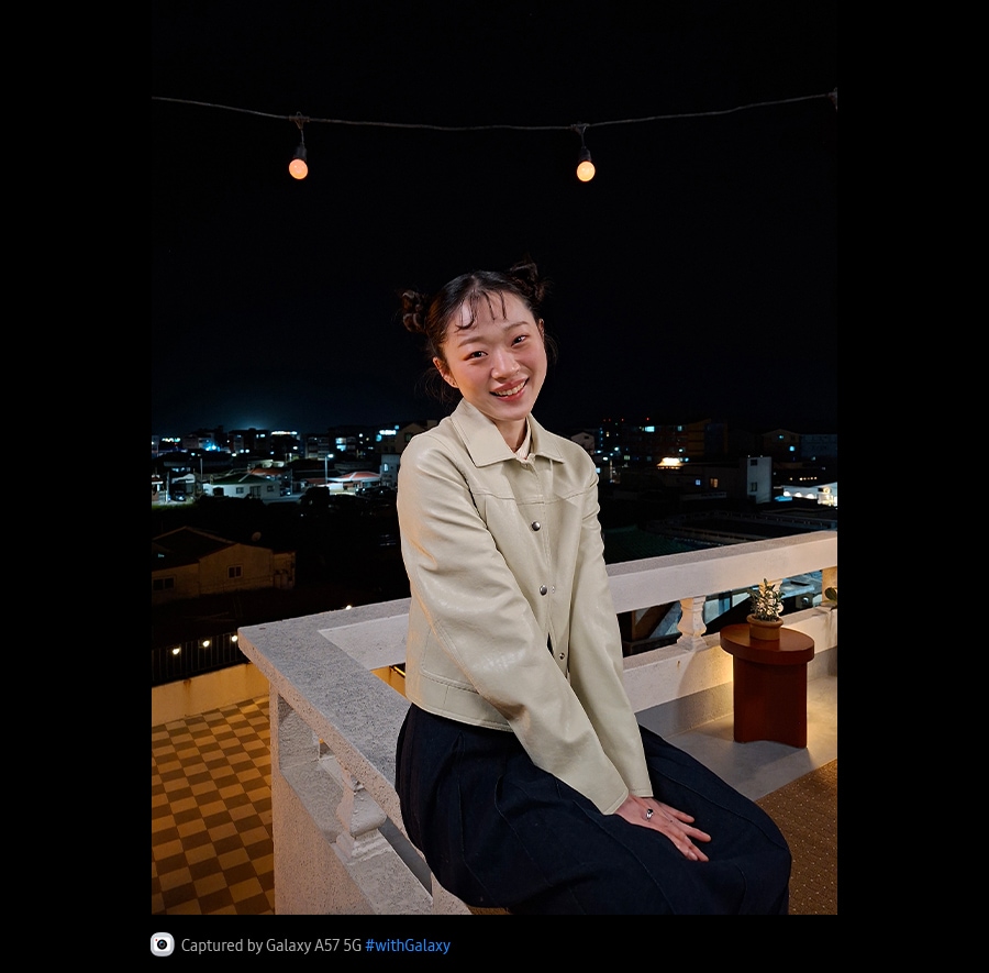 Photo of a young woman leaning on an outdoor terrace taken at night with Galaxy A57 5G. Photo details are bright and clear thanks to optimized Nightography.