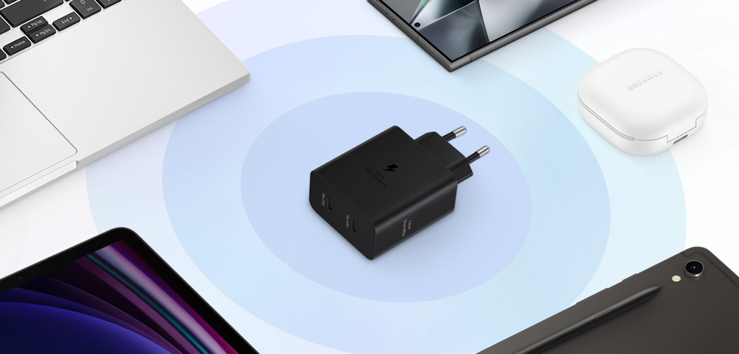 A imagem mostra uma vista superior de vários dispositivos Samsung Galaxy, com um adaptador de energia preto posicionado no centro. No sentido anti-horário, à esquerda do adaptador de energia, vemos um Galaxy Book 3, um Galaxy Tab S9 com parte da tela voltada para cima, outro Galaxy Tab S9 com a S Pen e parte da traseira voltada para cima. À direita, são exibidos os Galaxy Buds 2 Pro na cor branca e um Galaxy S24 Ultra.