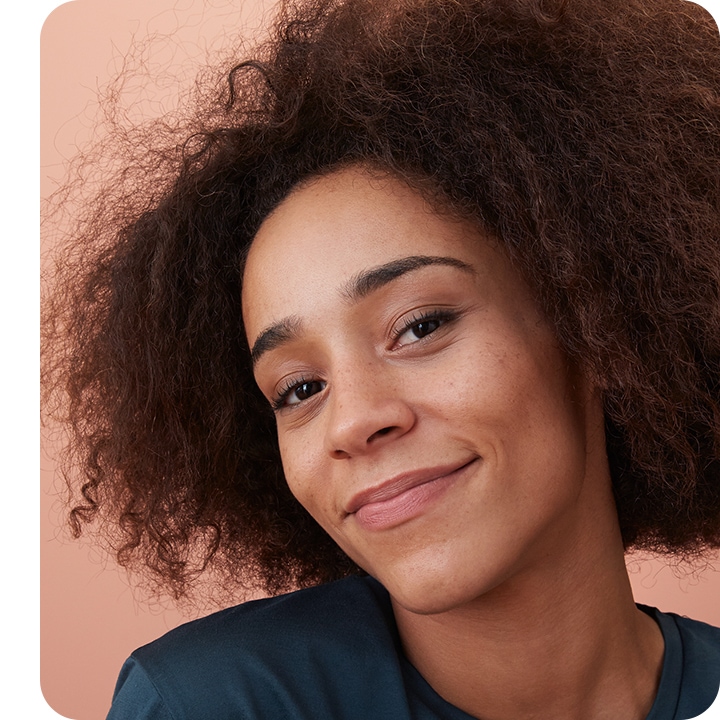 Selfie of a smiling young brown curly haired woman looking into camera lenses, with only a small part of background showing