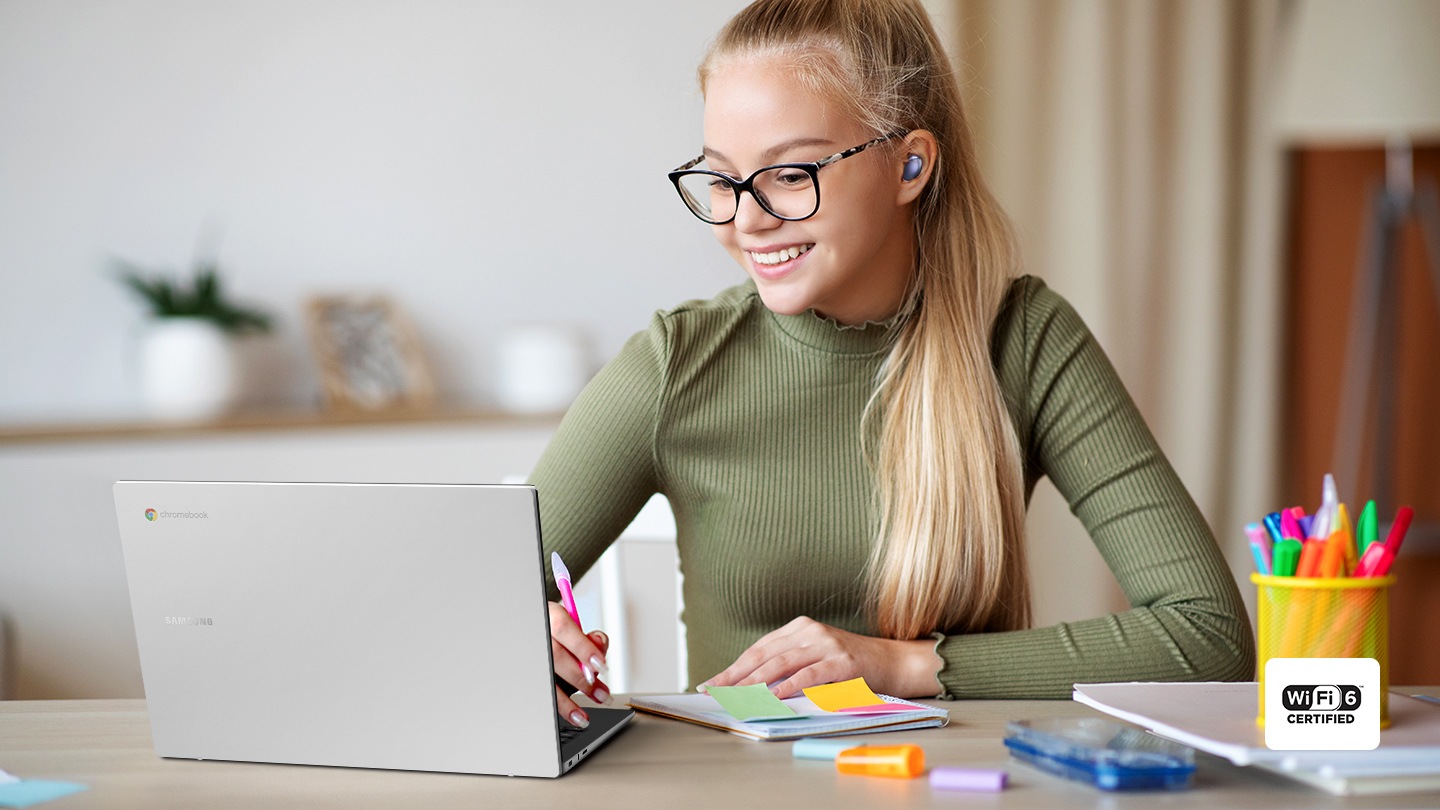 A person wearing Phantom Violet Galaxy Buds Pro is looking at the Galaxy Chromebook Go screen at a desk on which notebooks & pens are laid out. She has a pen in her hand. Wi-Fi 6 icon is shown.