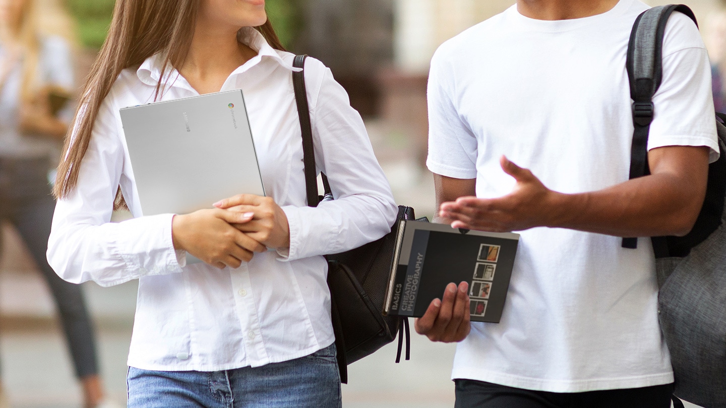 Two students are walking alongside each other. One of them is holding Galaxy Chromebook Go in her hands while the other one is carrying a textbook.
