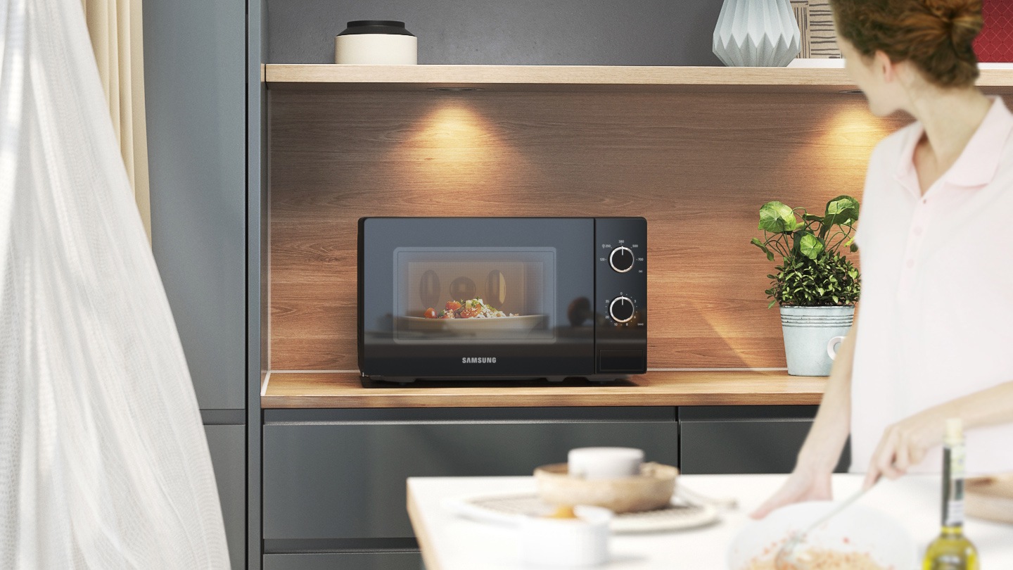 Mostra il forno a microonde su un piano di lavoro della cucina con una luce sopra. Una donna sta guardando il microonde e può vedere chiaramente cosa sta cucinando all'interno poiché la cavità del forno è illuminata brillantemente dalla sua illuminazione a LED.