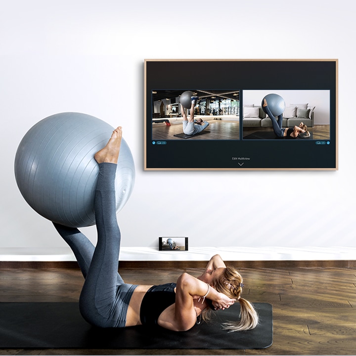  Una donna si sta allenando con una palla da ginnastica a terra con il suo smartphone accanto a lei. The Frame mostra cosa c'è sul suo smartphone e un video di un trainer sullo schermo della TV.