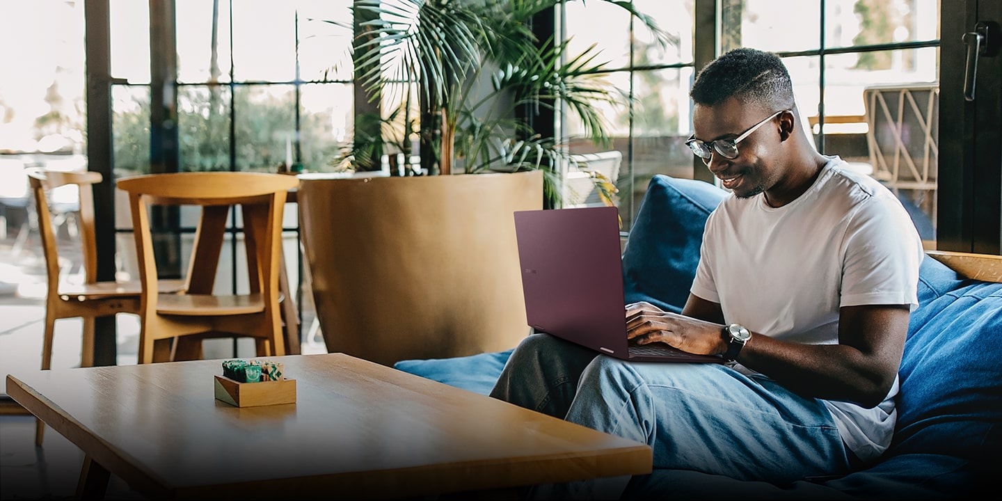 On the right is a man sitting on a large blue bean bag in a sunlit room with a large plant and wooden furniture surrounded by glass walls. The man is using a burgundy-colored Galaxy Book2 Pro 360 from his lap, smiling.