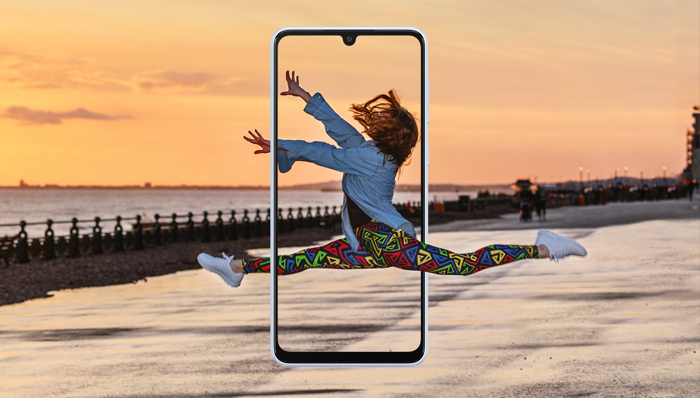 Una donna ripresa a metà di un salto che assomiglia a una figura di danza classica. Al centro di un paesaggio con un bellissimo tramonto, lo schermo di un Galaxy A33 5G incornicia la donna a metà del salto.