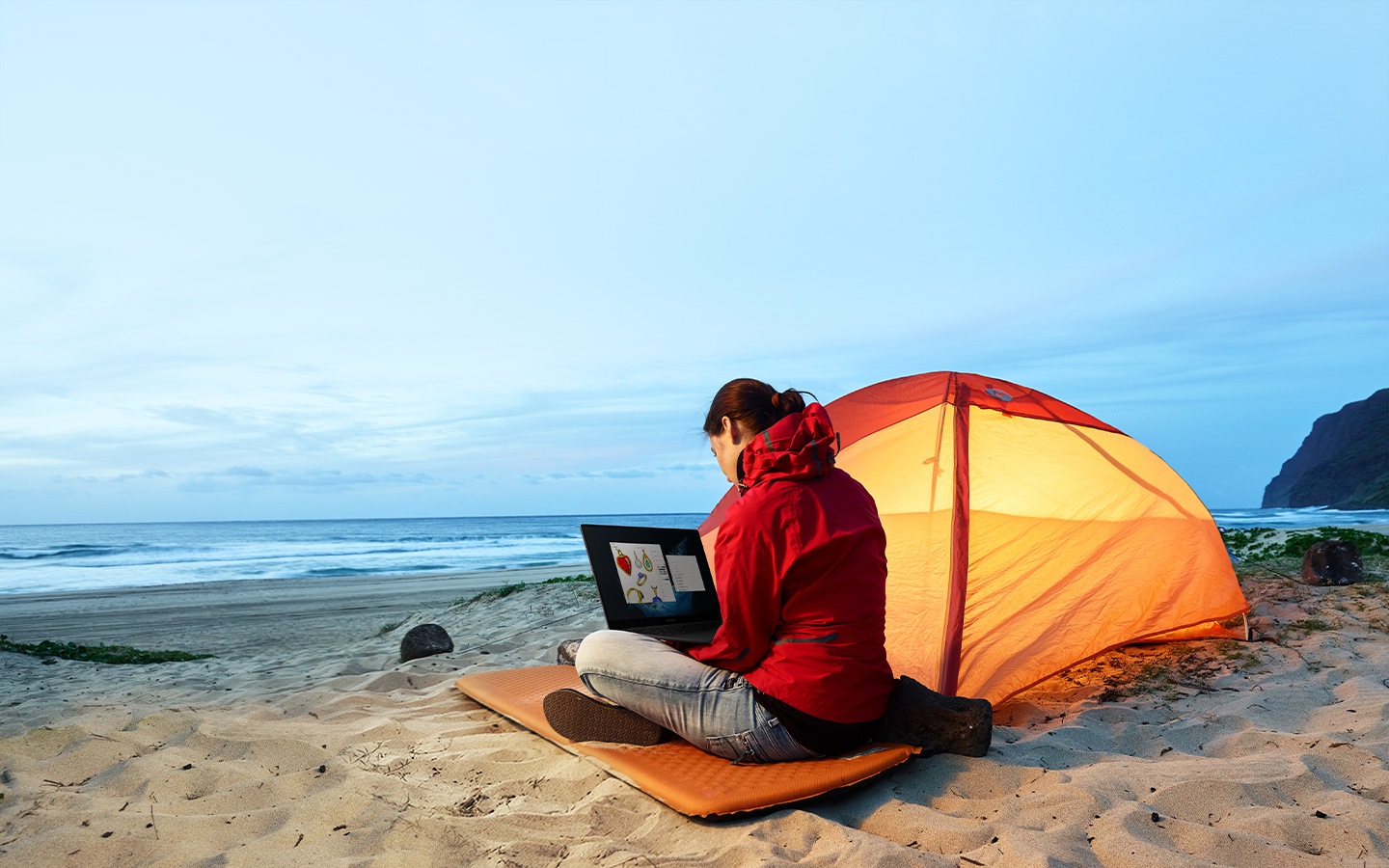A woman in jeans and a red windbreaker is sitting next to her orange tent on a beach. She is sending files using a Galaxy Book2 Pro 5G.