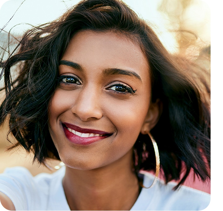 A portrait shot of a woman smiling, looking into the camera with a bright, yet blurry outdoor background shown behind her to highlight her face.