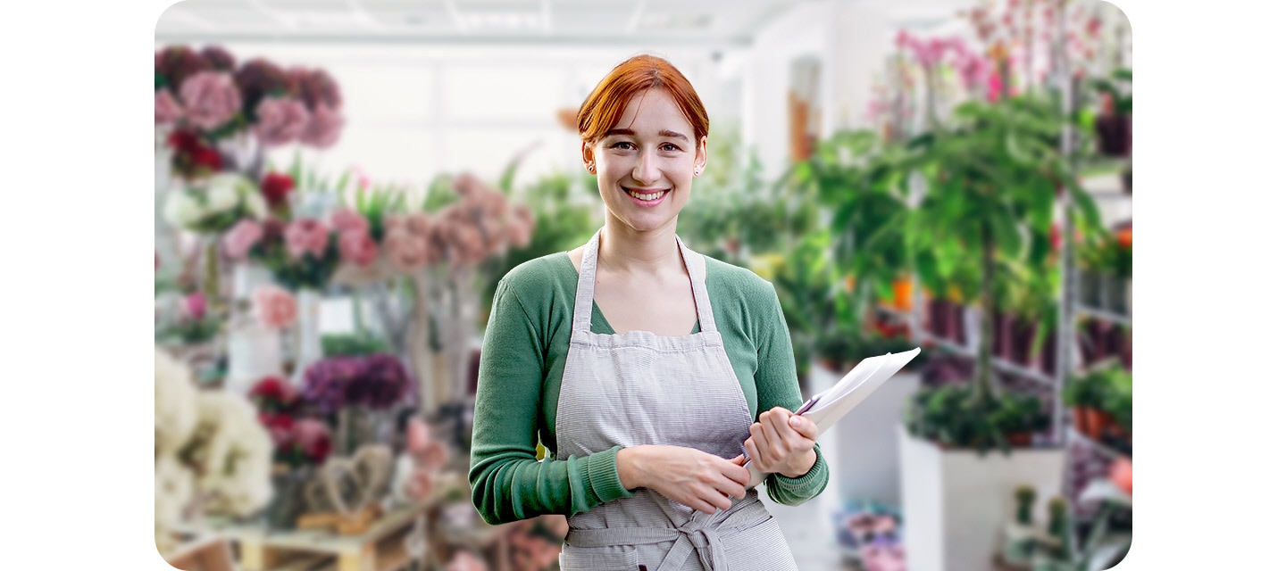 A woman is smiling and holding paper and pen in a flower shop. She stands out clearly against a blurred background.
