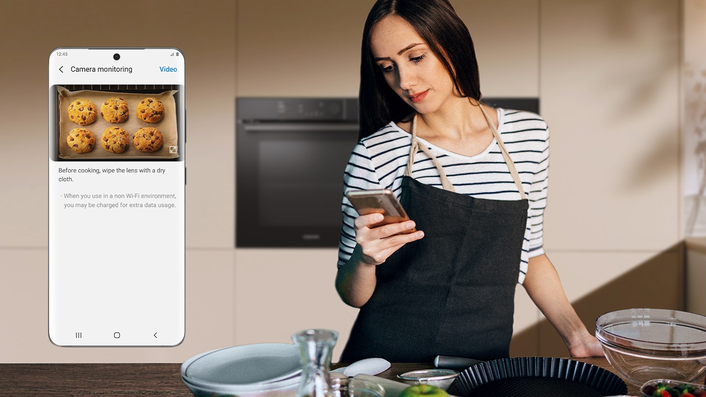 Mostra una donna davanti a un forno che guarda il suo smartphone. Un primo piano dello schermo del telefono mostra la funzione di monitoraggio della telecamera dell'app SmartThings, che mostra un video in tempo reale dei biscotti che vengono cotti nel forno.