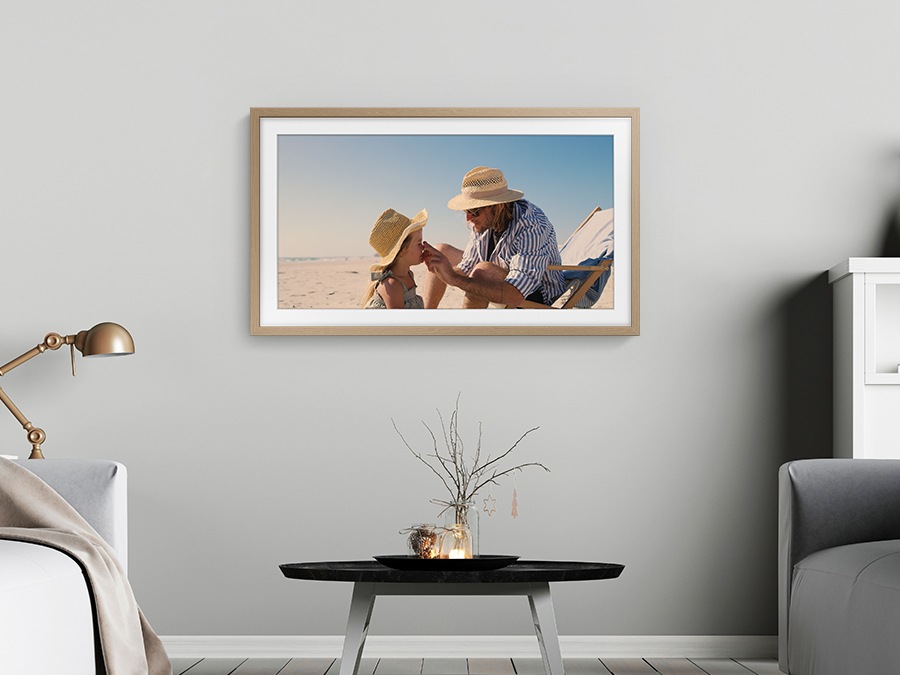 A father is touching his daughter's face on the beach. The moment is captured on The Frame, which is hanging on a wall in a room.