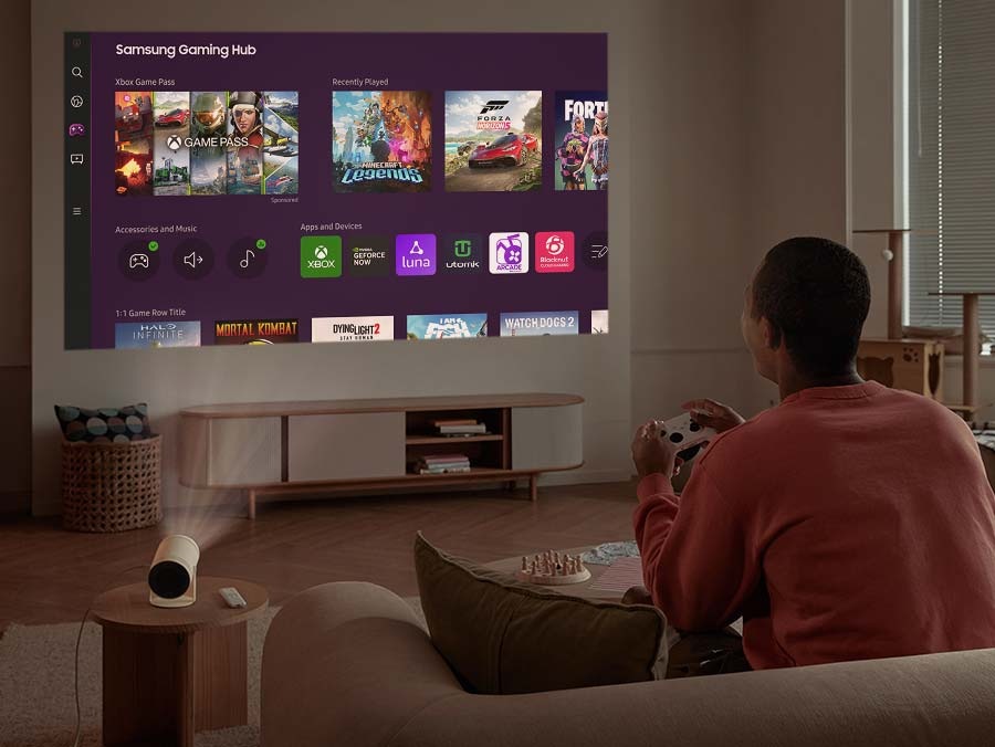 A man sits on a couch in his living room and The Freestyle is projecting a content onto the wall. Onscreen is the Gaming Hub GUI.