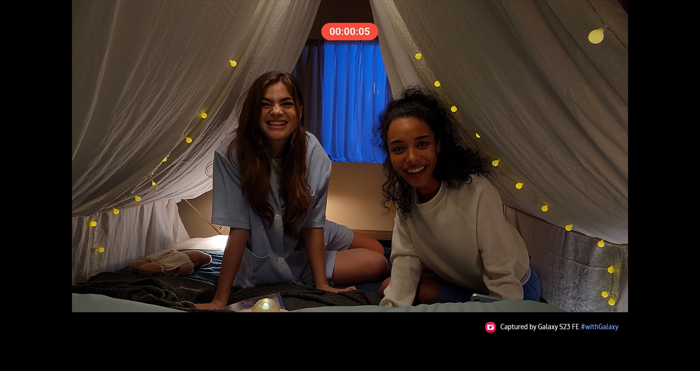 Due donne sorridenti salutano mentre entrano sotto una sorta di tenda drappeggiata con un tessuto beige mentre qualcuno le riprende con uno smartphone. La stanza è poco illuminata ma il dettaglio e il colore del video risultano nitidi e bilanciati, grazie alla tecnologia Nightography della fotocamera posteriore di Galaxy S23 FE. Scritta: Immagina scattata con Galaxy S23 FE #withGalaxy.