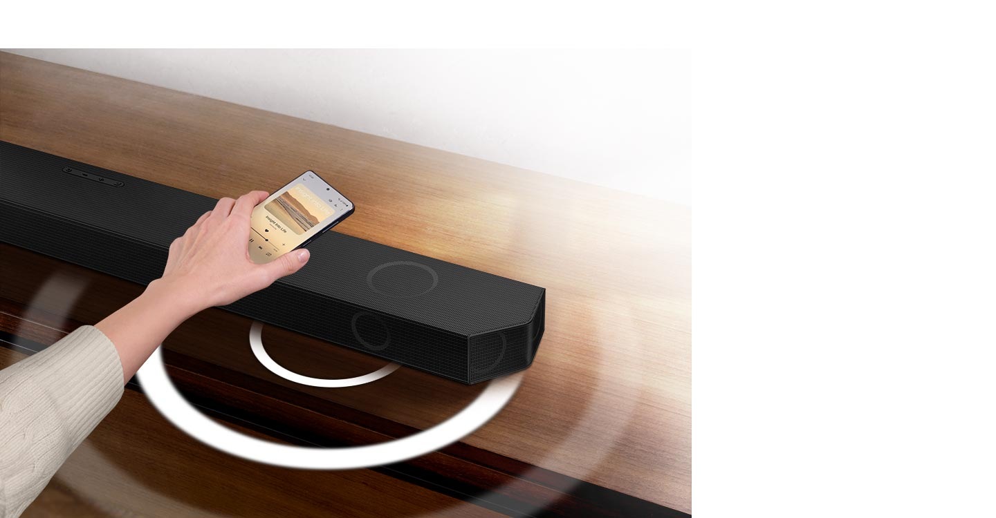 A hand holds a phone playing music and taps it lightly against a Soundbar. Instantly, the Soundbar gives off a series of round sound waves, indicating that it's playing the same music as the phone.