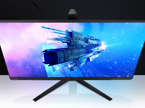 A Monitor which is seen from above shows large rectangular spaceship floating in space passing by another, larger space ship on its screen. The ship is surrounded by vibrant blue and purple star dust and countless stars. The front of the smaller spaceship is reflecting a bright light coming from an unknown source off screen.