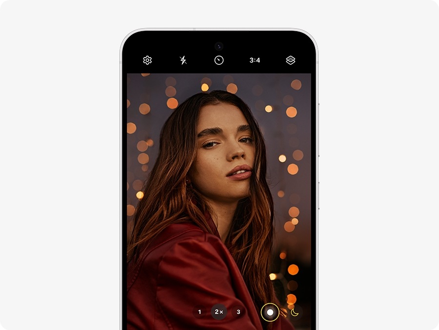 Galaxy S24 FEデバイスが正面から見え、画面には薄暗い部屋で微笑む女性の写真が映っている。画面では、操作中のカメラが2倍ズーム、3:4のアスペクト比に設定されている。