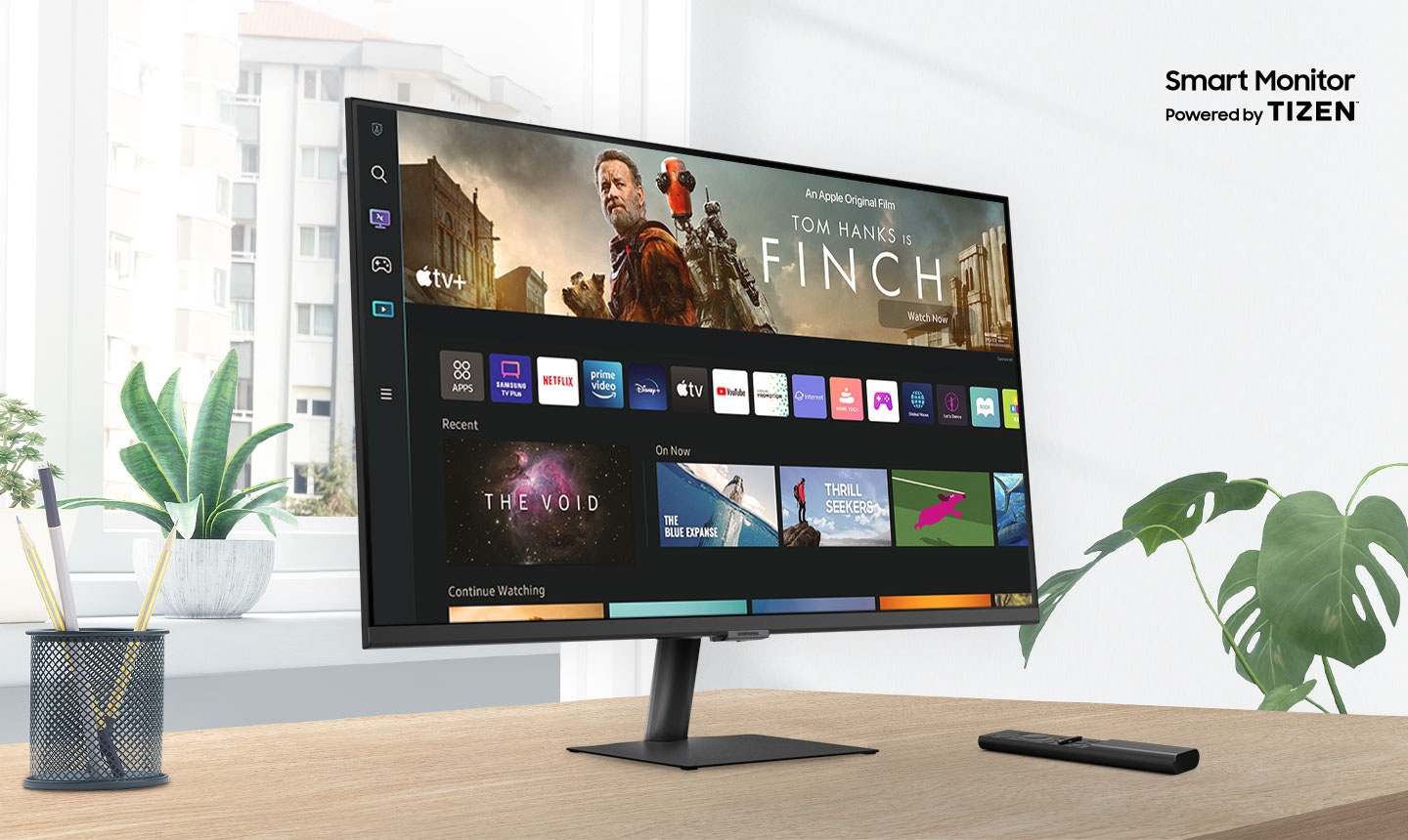 The monitor sits on top of a wooden desk in front of a window surrounded by plants.  The words †Smart Monitor Powered by Tizen' are shown above the monitor.  The screen shows a variety of content apps and individual content choices in a card format.  A remote rests next to the monitor.