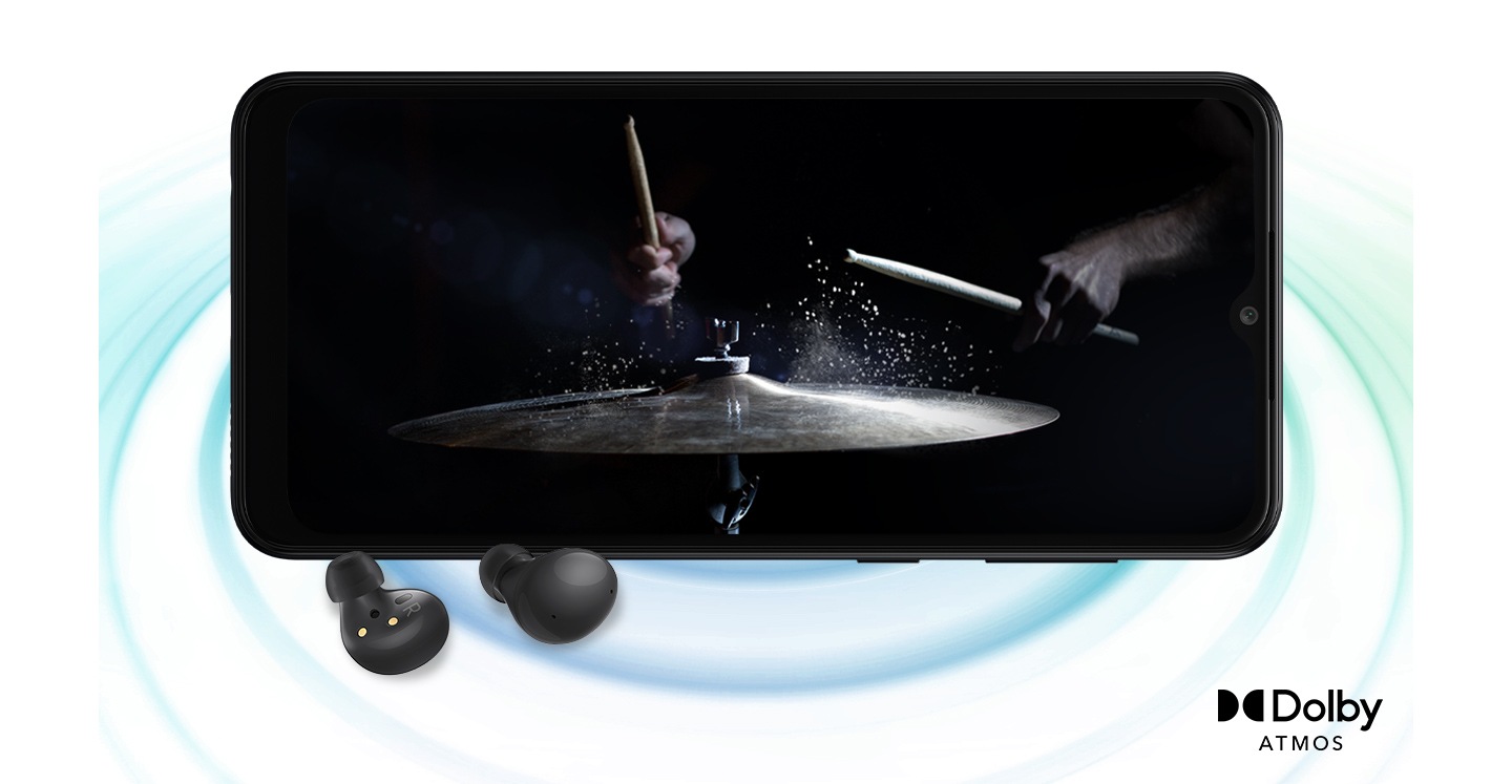 Galaxy A04 in landscape mode and an image with a person playing drums in the black background onscreen. A pair of black Galaxy Buds2 are placed in front of the device. On the right bottom is a logo for Dolby Atmos.