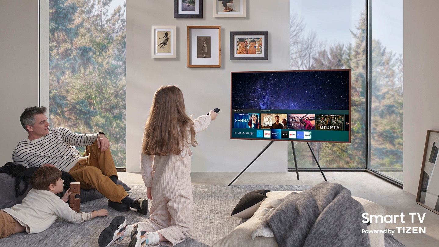 Un padre y sus dos hijos disfrutan de The Frame, que está sobre el soporte Studio. En la pantalla, se observa el menú de la interfaz de usuario de la Guía universal que la niña está controlando con One Remote. Se puede ver el logotipo de Smart TV Powered by Tizen.