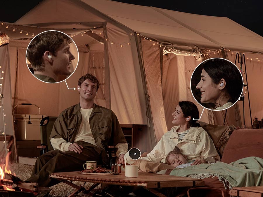 Una pareja está acampando afuera y The Freestyle está en marcha. Están escuchando el mismo audio en Galaxy Buds separados. Un bebé duerme en el regazo de la mujer.