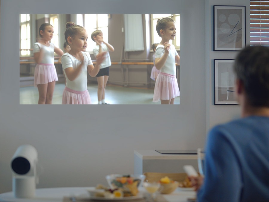 Un hombre está reproduciendo un video de niños en una clase de ballet con The Freestyle. El video está ligeramente obstruido por marcos que cuelgan de la pared, pero The Freestyle lo detecta y aleja automáticamente la proyección de los marcos.