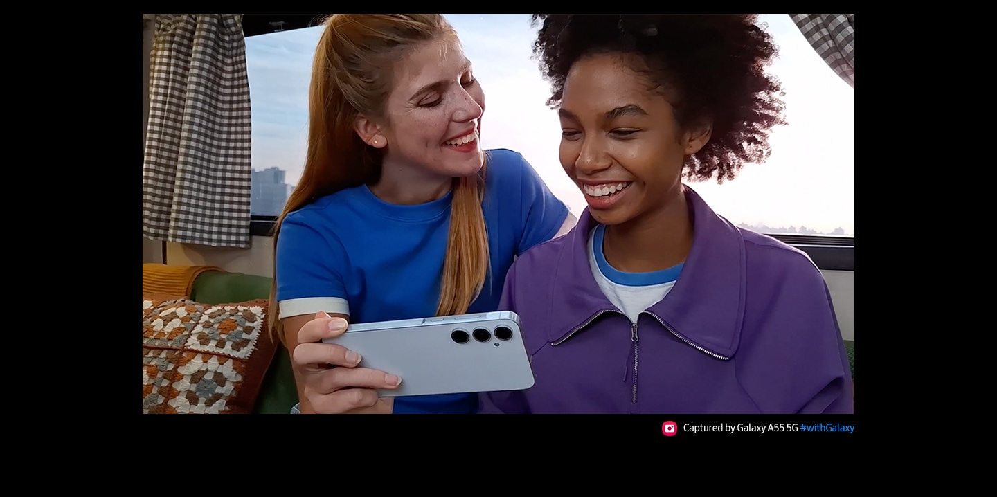Un video de dos amigos mira teléfonos inteligentes. Están sentados juntos, sonriendo con una vista panorámica de la ciudad a través de la ventana que está detrás de ellos. El texto dice Capturado con el Galaxy A55 5G #withGalaxy.