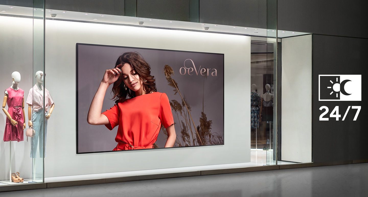 The image shows the front of a store with a large digital screen in the window. The screen displays an ad for the "oeVera" brand, featuring a woman in an orange dress. A "24/7" icon is visible to the right, indicating the display is designed for continuous operation.