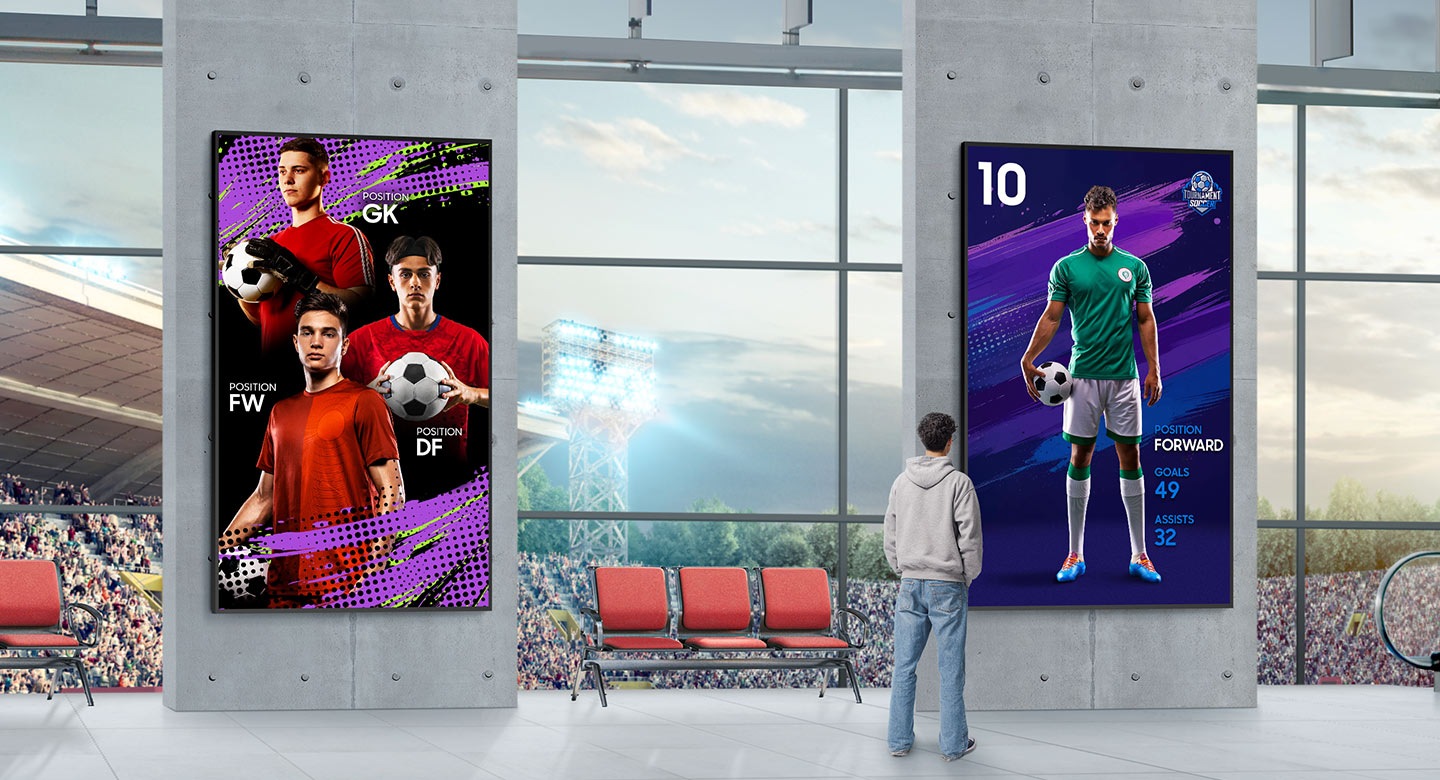 The image shows the inside of a stadium with a large window looking out onto the field. Two large, vertical digital displays are mounted on concrete pillars. The screen on the left shows three soccer players, while the one on the right shows a single player with his stats. A person is standing in front of the right screen, looking at the display.