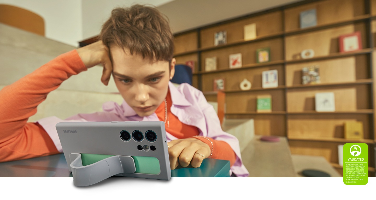 A young student is resting her head on her hand while looking at the screen of her Galaxy S24 Ultra in a taupe Standing Grip Case. The Strap forms a stand that helps the phone stand on its own, demonstrating practical situations by securely fixing the strap for comfortable screen viewing. The text reads UL VALIDATED STANDING GRIP CASE (EF-GS928) CONTAINS A MINIMUM OF 22% POST-CONSUMER RECYCLED CONTENT, CONSISTING OF 41% POST-CONSUMER RECYCLED CONTENT IN THE PLASTIC OF STANDING GRIP CASE. UL.COM/ECV