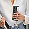 Una mujer joven vestida semiformal camina al aire libre, sosteniendo un dispositivo Galaxy Z Fold5 cubierto con un estuche con correa y sus dedos deslizados debajo de una correa de color rojo.  En la parte inferior del estuche se encuentran accesorios coloridos.