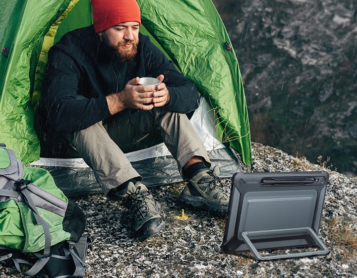 Un hombre que sostiene una taza está sentado frente a una tienda de campaña sobre una superficie rocosa, mirando un dispositivo Galaxy Tab S9 con una cubierta para exteriores colocada horizontalmente usando el pie de apoyo en la parte posterior.