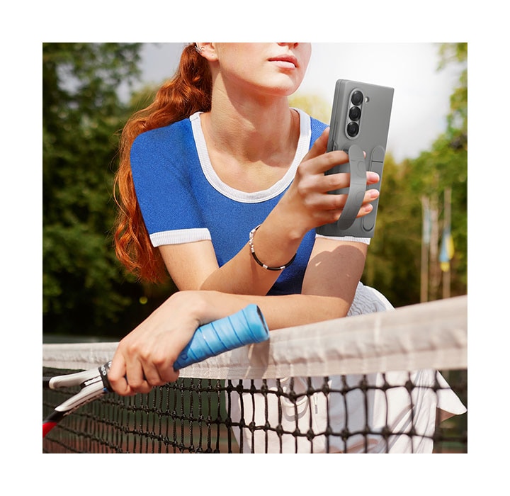 A young woman at a tennis court is leaning on the tennis court net, holding a Galaxy Z Fold6, comfortably. The phone is in a Silicone Case in gray and the woman holds the phone with the case grip looped through her fingers.