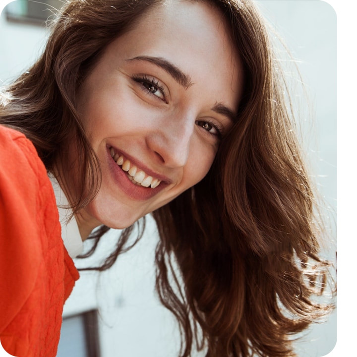 Selfie d'une jeune femme aux cheveux bruns souriante à la recherche dans les objectifs de l'appareil photo, avec seulement une petite partie de l'arrière-plan flou montrant