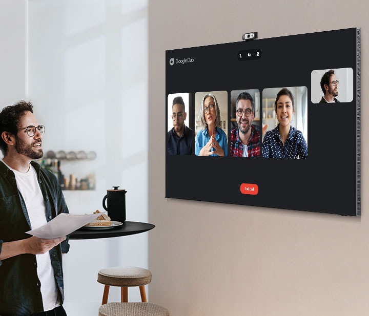 A man uses Neo QLED's built-in Video Call with Google Duo app to participate in a teamwork meeting from a home dining room. TV onscreen is showing four coworkers.