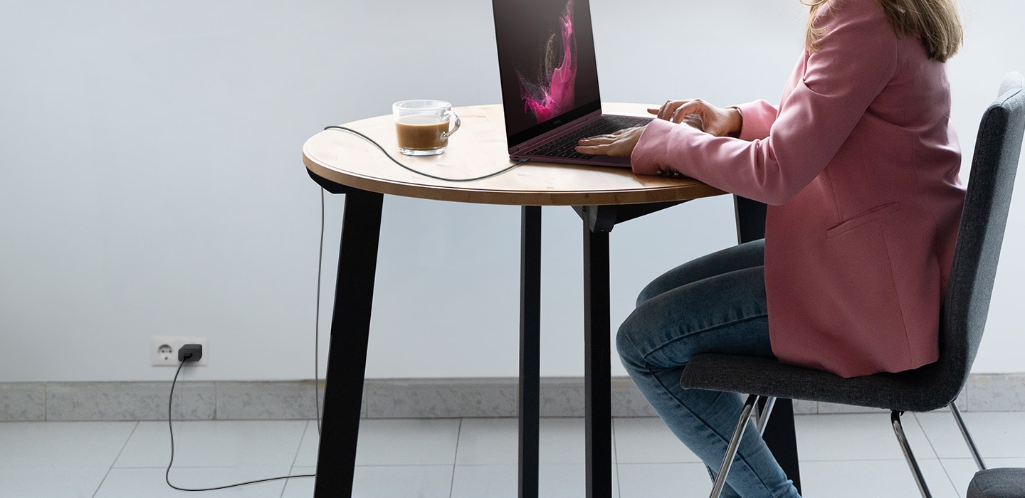 A person sitting at a table and using a Galaxy Book laptop. The USB cable is plugged in to the laptop, with the other side plugged into the wall outlet with an adapter. There is slack in the cable because of its long length.