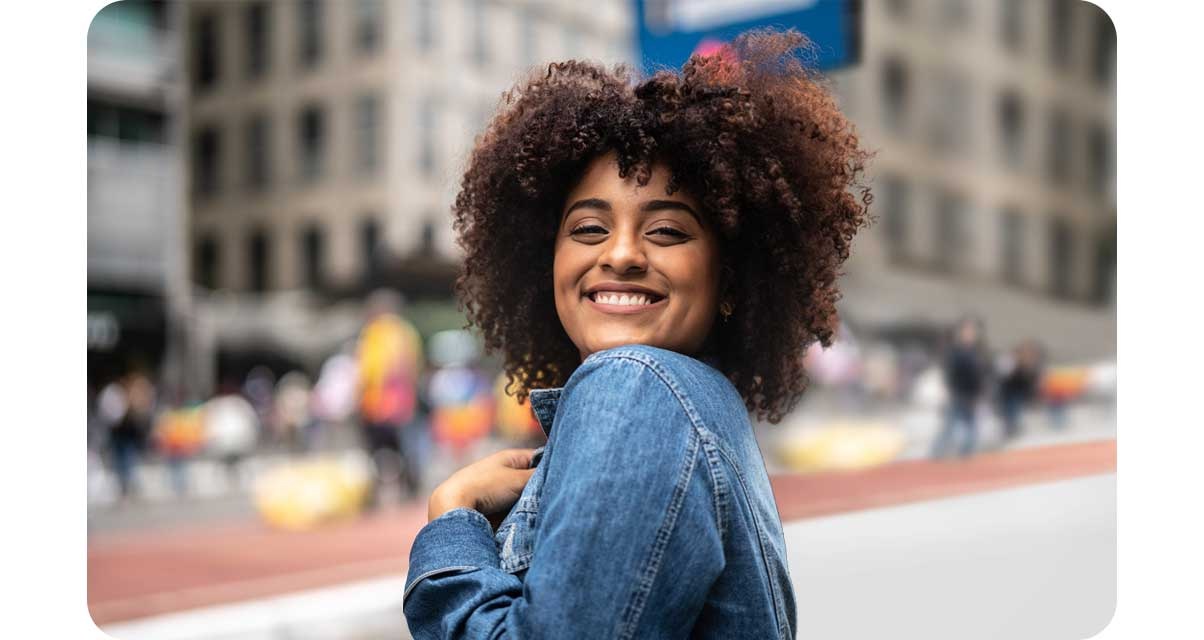 A smiling woman is shown in the center of an urban setting with the background, showing buildings and people, blurred behind her.