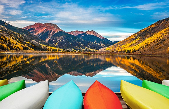 Une belle photo de paysage avec un ciel bleu et des montagnes en arrière-plan, un lac immobile et réfléchissant et des canoës colorés au premier plan.