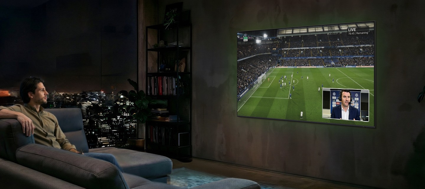 Man watches football on Samsung TV at night. Soundwaves emanate from TV's side and front. He speaks into remote control ‘Bixby, AI Football Mode ON’. AI Football Mode turns on, soundwaves show balanced crowd and commentator sound, and the field looks more crisp and vibrant than when it's off.