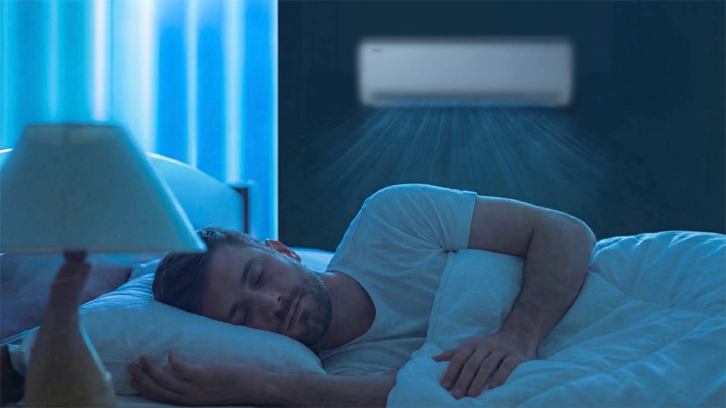In the dark bedroom, a man lies asleep. A wall-mounted air conditioner operates on the back wall, gently releasing cool air downward. The bedside lamp is off, and the soft blue light seeping through the window adds to the room's tranquil atmosphere.