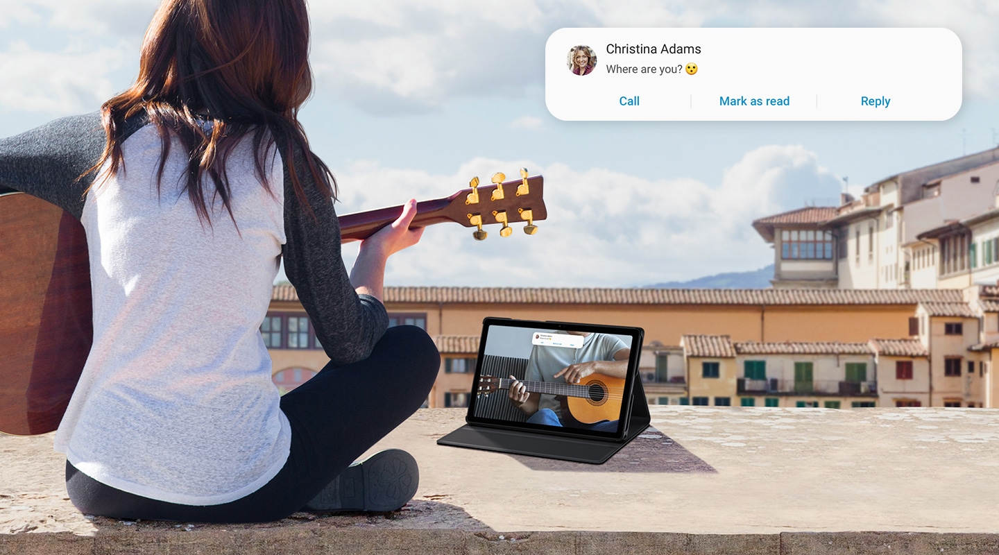 A woman sits with her guitar, watching a guitar tutorial on her Galaxy Tab A7. A text from her friend appears at the top.