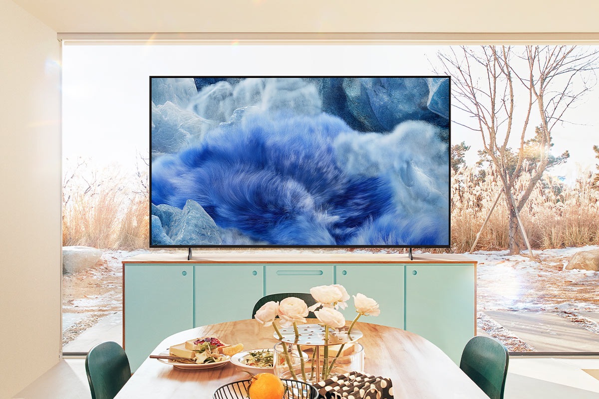 A QLED TV in a kitchen area displays a blue grassy landscape. Behind the TV, a glass window reveals the outdoor scenery.