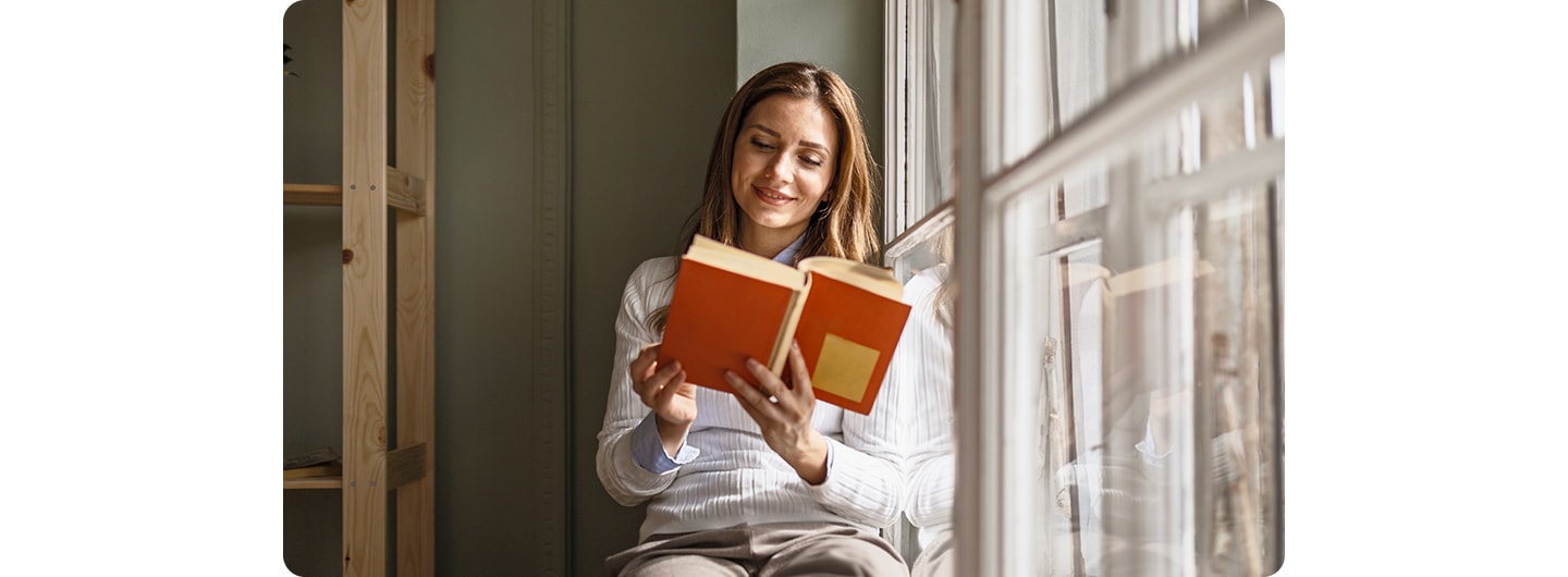 A woman sitting indoors, reading by a window. A line down the screen splits it into two sections, comparing a low-light shot on the left with a bright-light shot on the right.