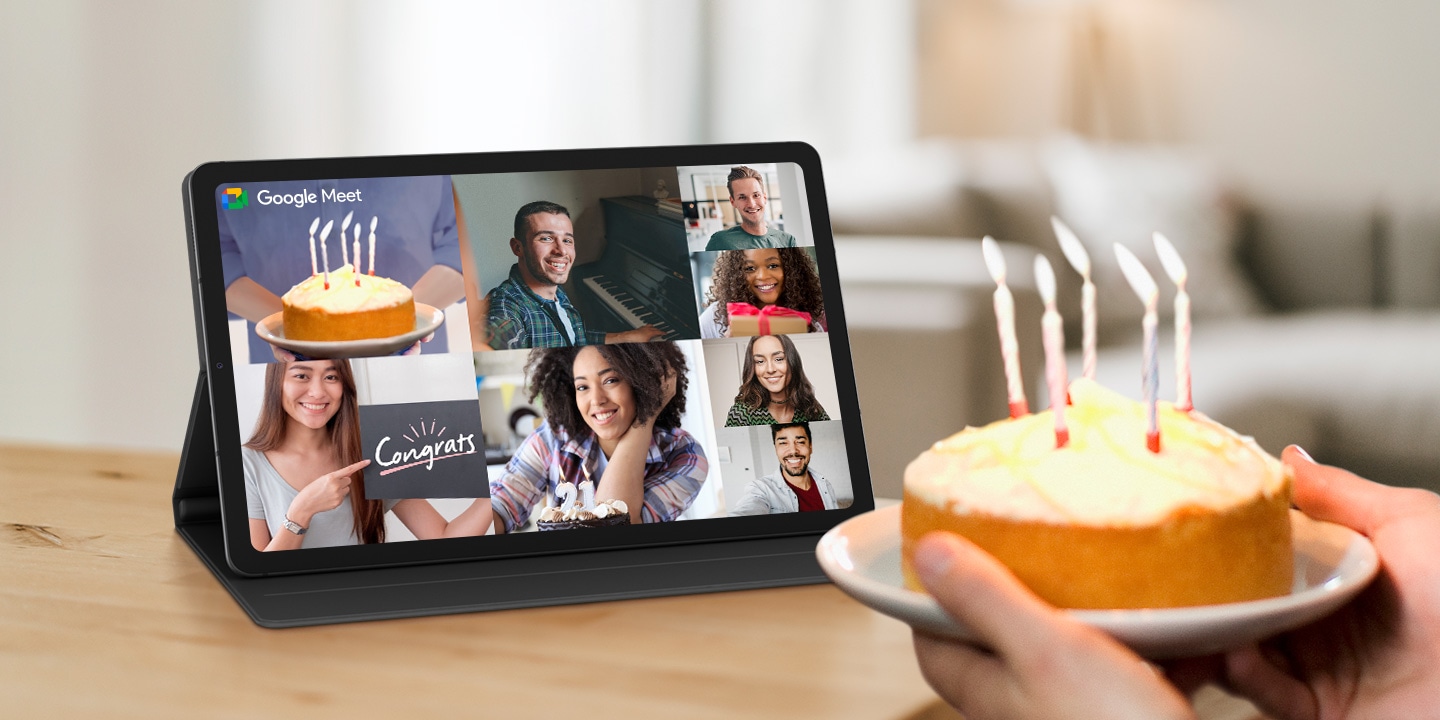 A person is holding a cake with lit candles in front of a Galaxy Tab S6 Lite device, fixed in landscape orientation using the Book Cover. The screen of the device is divided into rectangular images to show that several people are connected through video calling. In one image, the cake with lit candles is shown, while in each of the other images there is a person smiling. The Google Meet logo is displayed on the top left side of the screen.