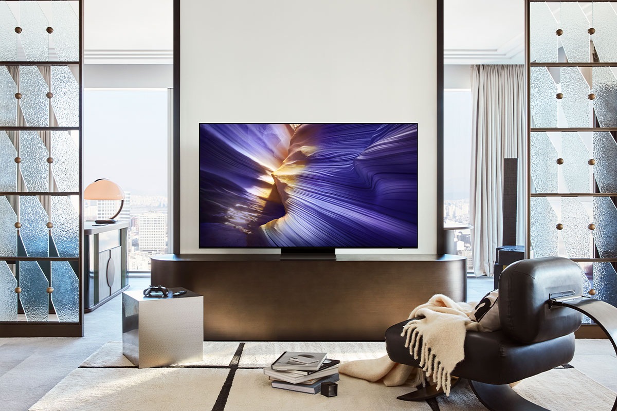 A Samsung OLED TV is set on a stand in a living room. The TV screen displays an abstract purple landscape.