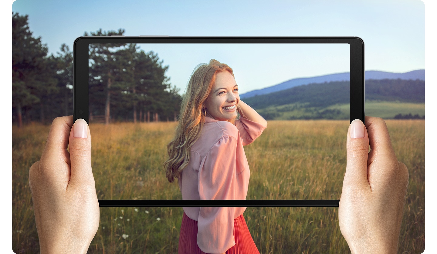 Hands holding Galaxy A7 Lite. Onscreen is a woman standing in a field. The photo goes beyond the boundaries of the screen to represent the wide display.
