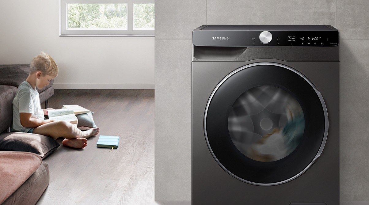 The boy reads the book during the day in the living room, while the washer runs silently next to him.