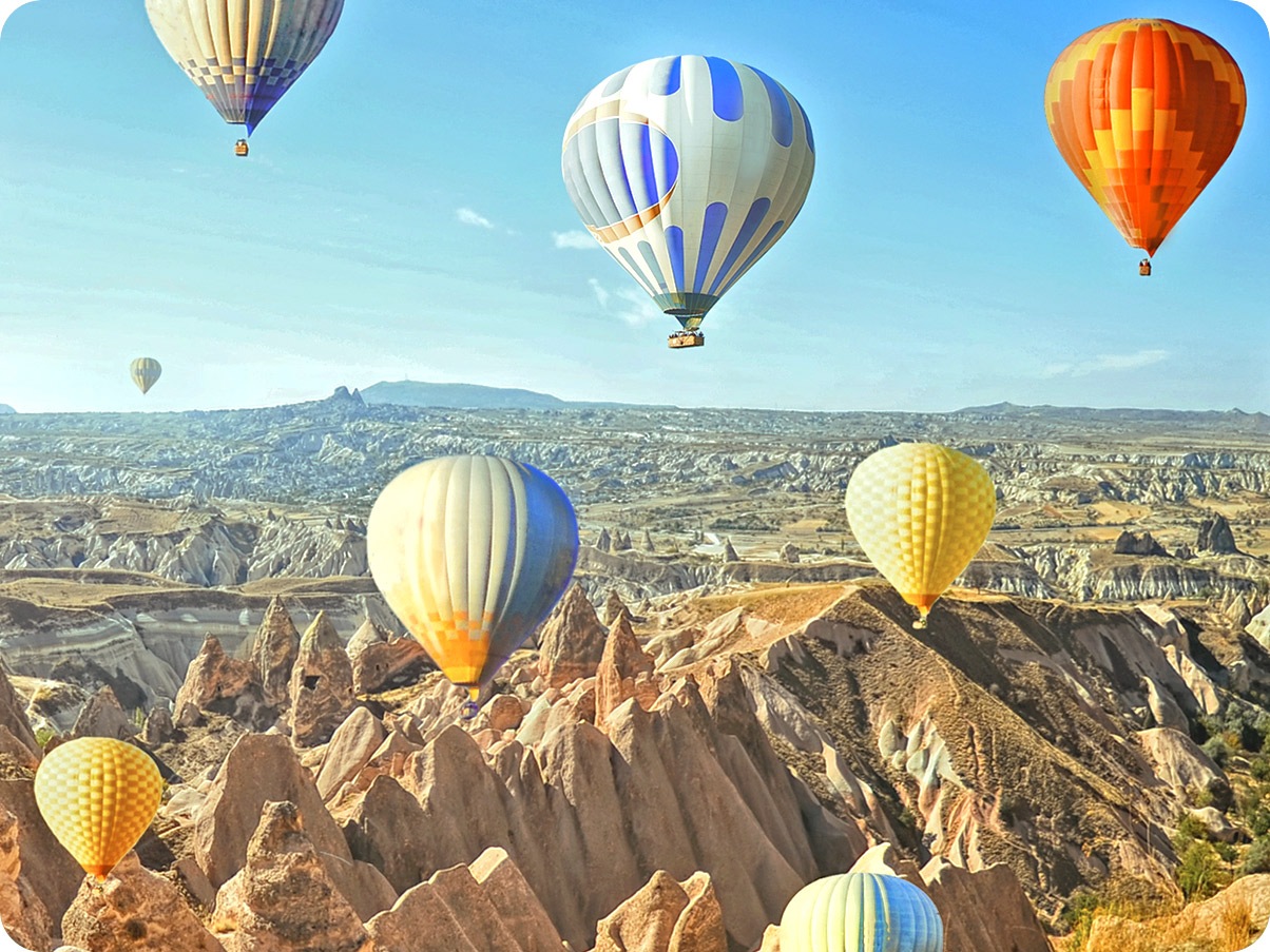 Foto tomada con cámara gran angular muestra coloridos globos de aire en el cielo y un vasto paisaje en el fondo.